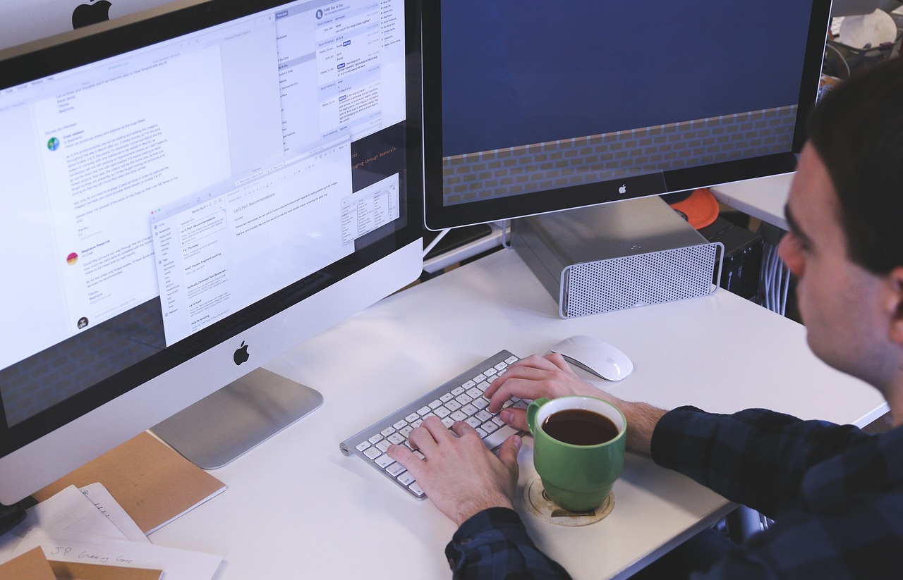 A man seated at a desk, engaged with two computer monitors, illustrating a modern work environment.