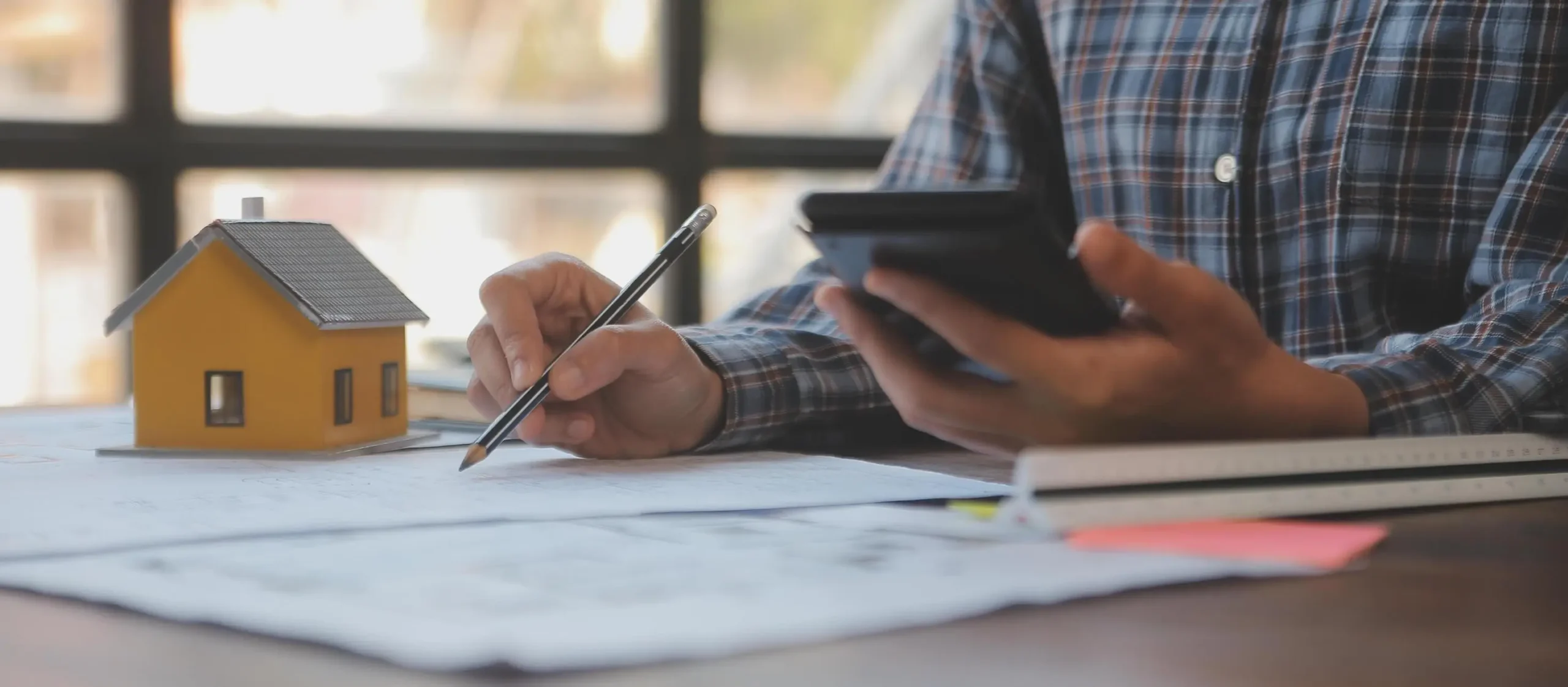 The image depicts a property valuer working with a model house and documents, while using a calculator.